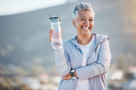 Mulher sorridente ao ar livre, segurando uma garrafa de água e vestindo roupa esportiva, representando bem-estar e os benefícios do uso de suplemento alimentar.
