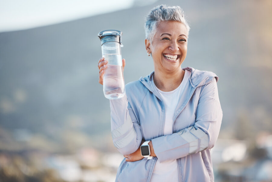 Mulher sorridente ao ar livre, segurando uma garrafa de água e vestindo roupa esportiva, representando bem-estar e os benefícios do uso de suplemento alimentar.